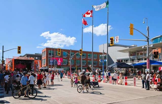 Crowds and cyclists fill a sunny downtown square with Canadian and Ontario flags, a stage with a screen, and Canada banners.
