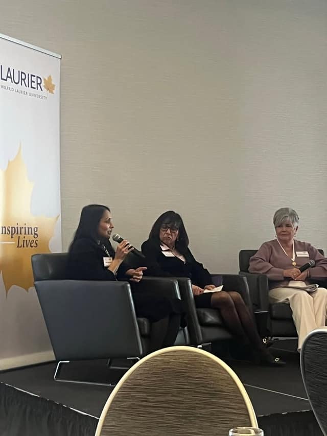 Three women sit in armchairs on a stage beside a Laurier "Inspiring Lives" banner; one speaks into a microphone.