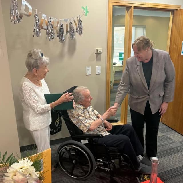 Norma holds a green folder as a woman in a gray blazer shakes hands with Harold, seated in a wheelchair, beneath a PARTY banner.