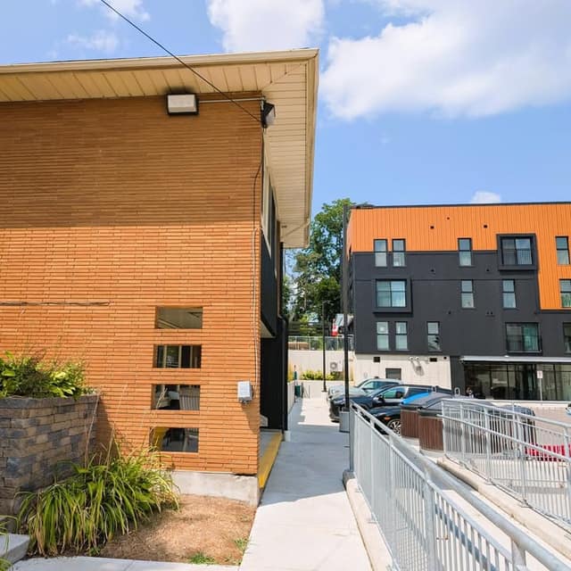 Orange brick building beside a walkway with a metal railing, and a black and orange apartment building with parked cars under a blue sky.