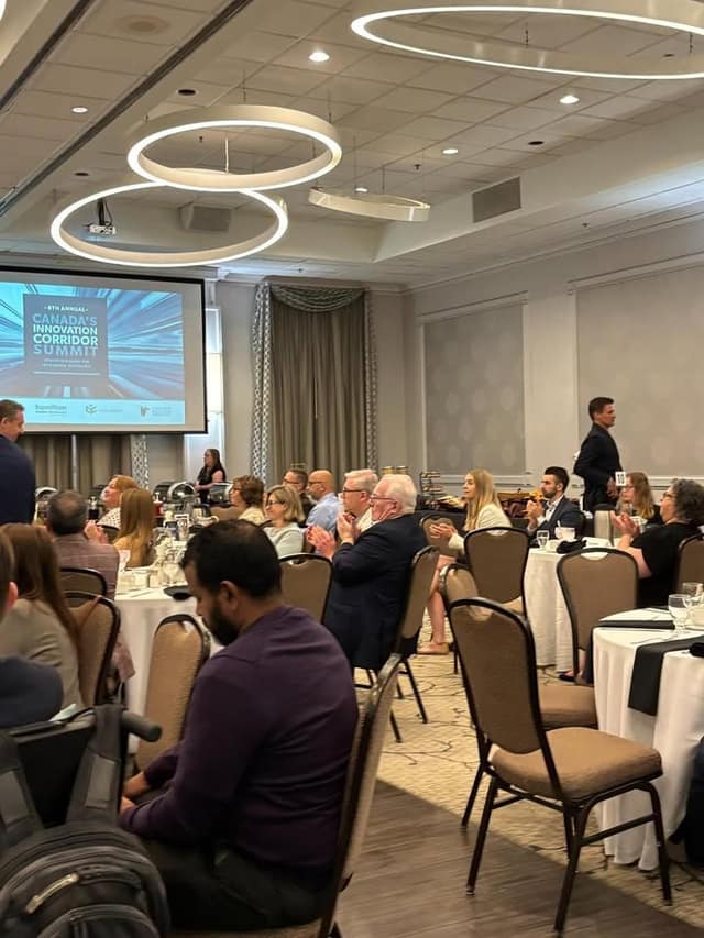 Attendees seated at round tables applaud in a banquet room, with a screen reading Canada's Innovation Corridor Summit at left.