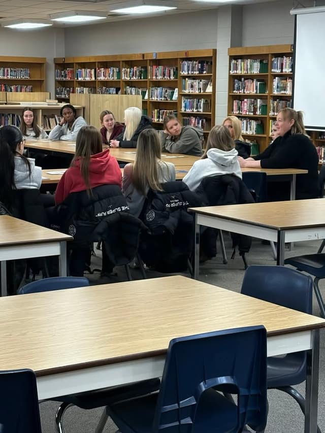 Students sit at wooden tables in a school library, listening to a woman in black seated at the table on the right.