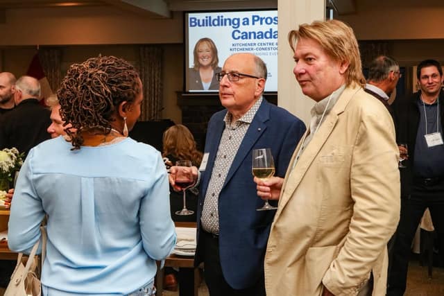 Three guests hold wine glasses and chat at a reception, with a projected screen reading "Building a Prosperous Canada" behind them.