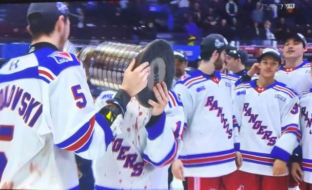 Kitchener Rangers players in white jerseys stand on the ice as one player drinks from the championship trophy's bowl.