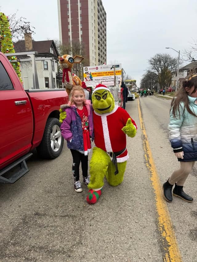 A girl in a purple jacket poses with a person in a Grinch costume on a parade route, with a red truck and a toy drive sign nearby.