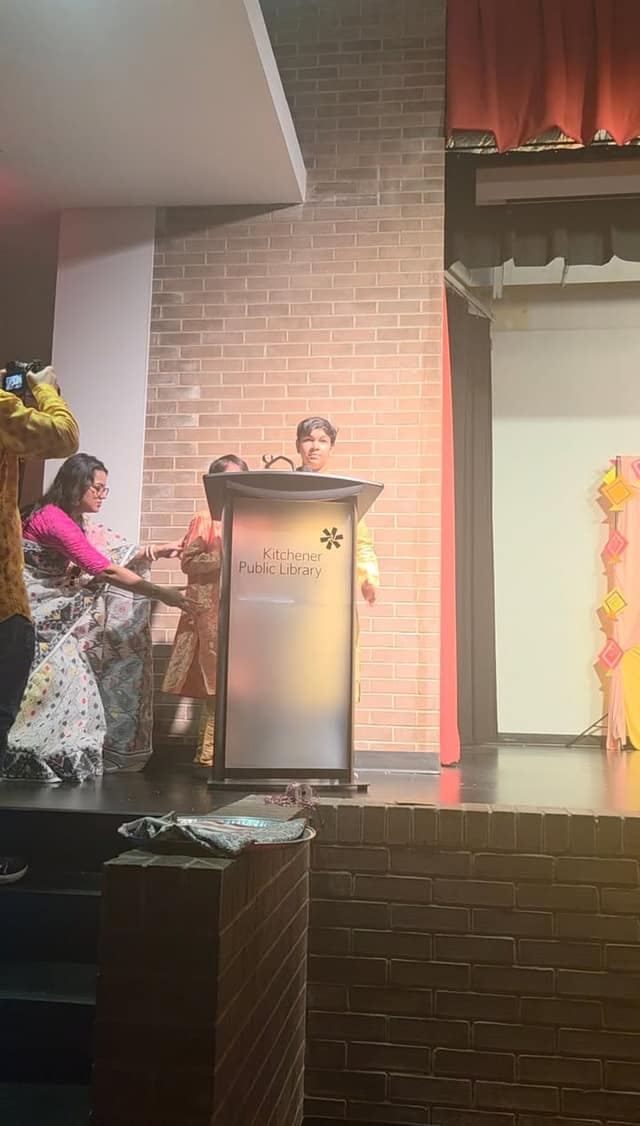 A child stands at a podium marked Kitchener Public Library on stage while a woman in a pink top adjusts a patterned sari nearby.