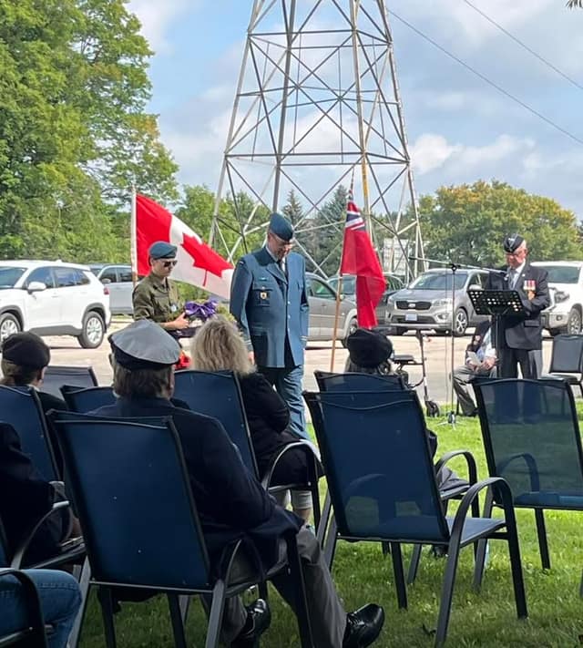 Guests in lawn chairs face a ceremony where a cadet holds a wreath near Canadian and Ontario flags and a speaker at a lectern.