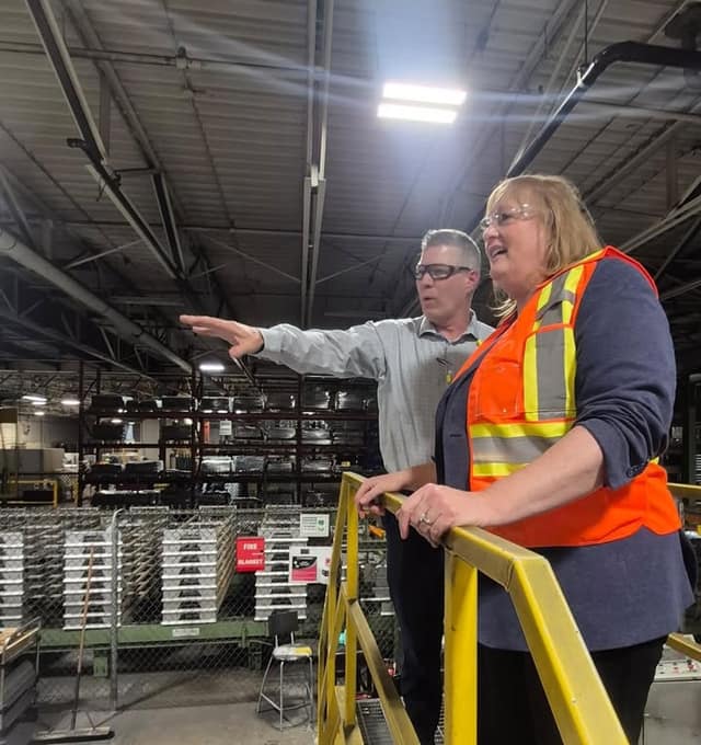 A man in glasses points across a factory floor while a smiling woman in an orange safety vest stands beside him at a yellow railing.