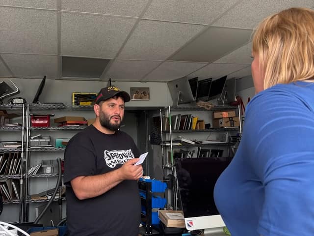 A bearded man in a cap and Speedway Trailers shirt holds a card and talks with a woman in blue, in a shop with shelves and monitors.