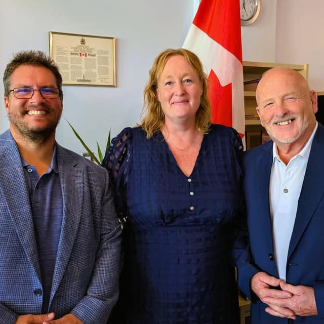 Two men and a woman in a navy dress smile together in an office, with a Canadian flag and a framed Charter of Rights behind them.