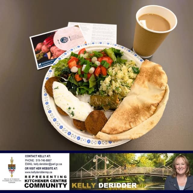 A paper plate holds falafel, salad, stuffed grape leaf, potato salad and pita, beside a cup of coffee and an information card.