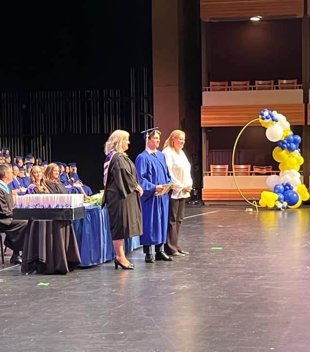 A graduate in a blue gown and cap holds a folder on stage between two women, beside a blue and yellow balloon arch.