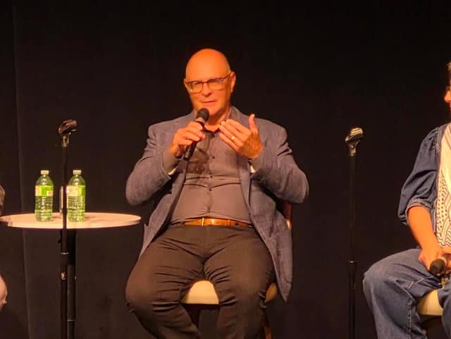 A bald man with glasses and a gray blazer speaks into a microphone and gestures with his hands beside a small table with water bottles.