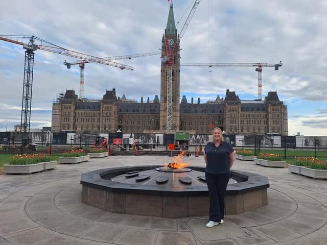 A woman in a dark top and pants stands beside the Centennial Flame on Parliament Hill, with cranes and scaffolding around the Centre Block.