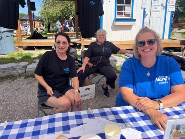 Three smiling women in Saint Mary's Kitchen t-shirts sit at a blue gingham-covered table outside a small white building.