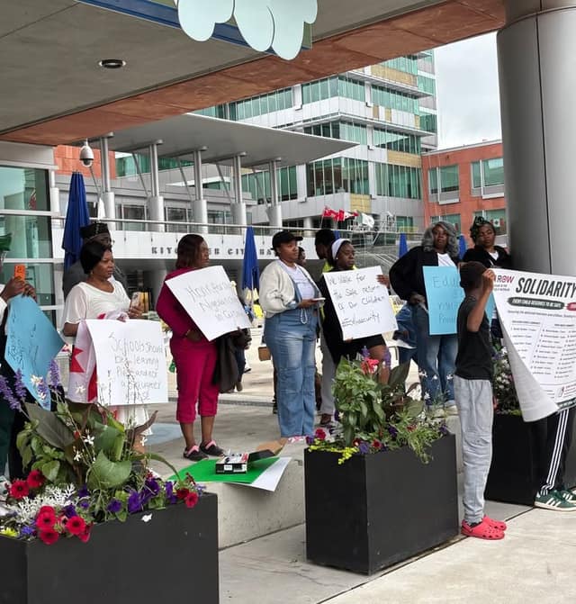 Vigil attendees hold handwritten signs beside flower planters outside Kitchener City Hall, with a large banner reading "Solidarity" at right.