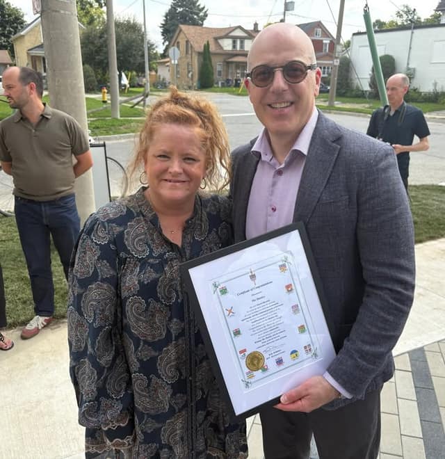 A woman and a man in tinted glasses hold a framed Certificate of Congratulations outdoors on a sidewalk, with houses behind them.