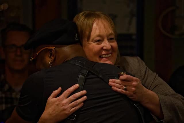 A woman in a tweed blazer smiles as she hugs a person in a black cap and black top, with a man watching in the background.