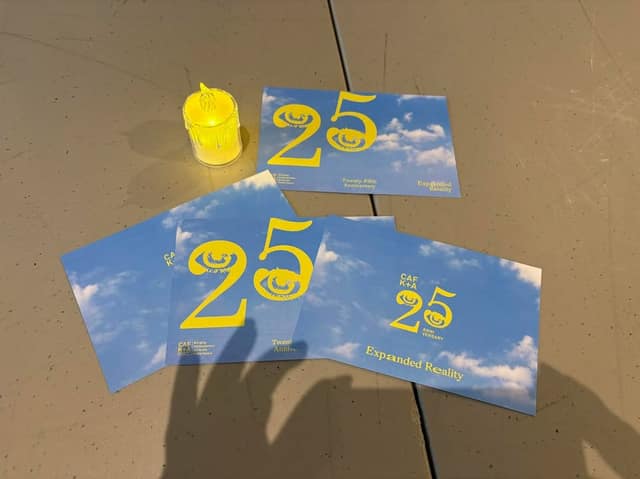 Four blue postcards marked 25, Twenty-Fifth Anniversary and Expanded Reality lie on a gray table beside a small yellow flameless candle.