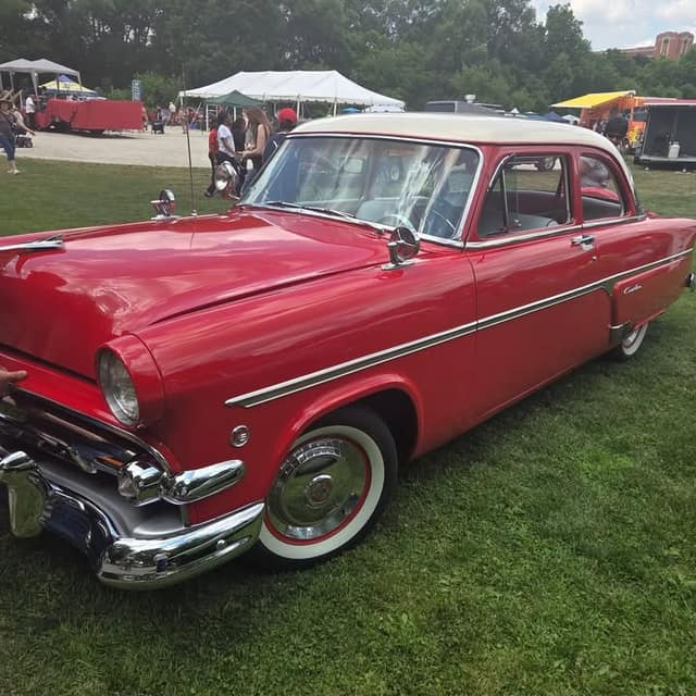 A restored red 1950s car with a white roof and whitewall tires is parked on grass at an outdoor show, with tents behind it.