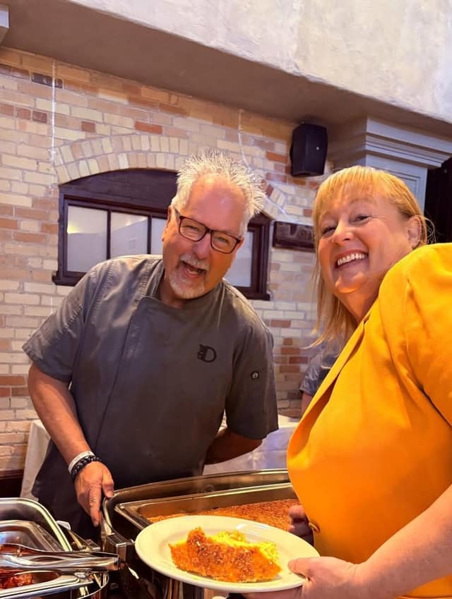 A smiling chef in a gray jacket stands at a buffet tray beside a laughing woman in a yellow blazer holding a plate of food.