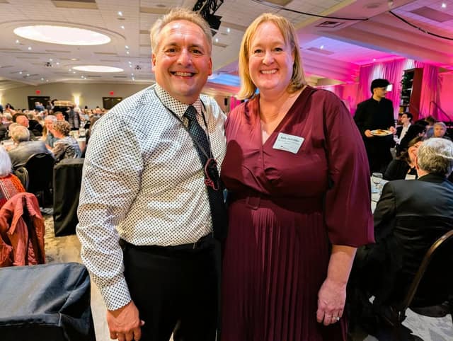 A smiling man in a dotted shirt and tie stands beside a smiling woman in a burgundy dress wearing a name tag, in a banquet hall.