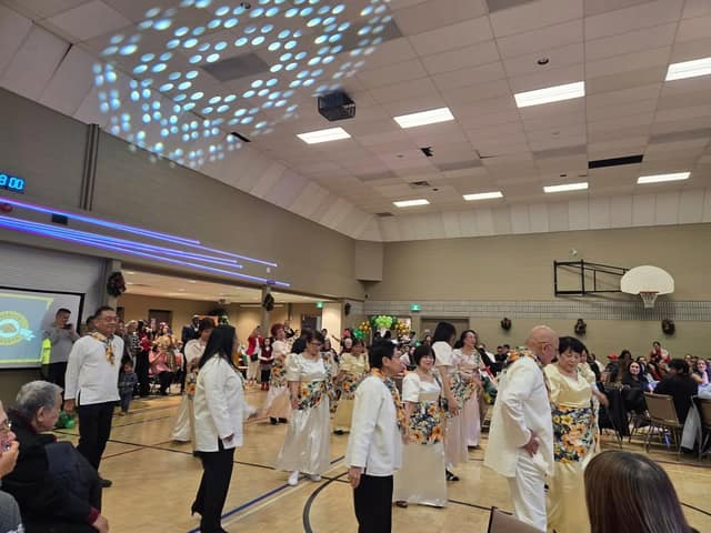 Dancers in white and floral Filipino formal wear perform on a gym floor while seated guests watch under patterned lights.
