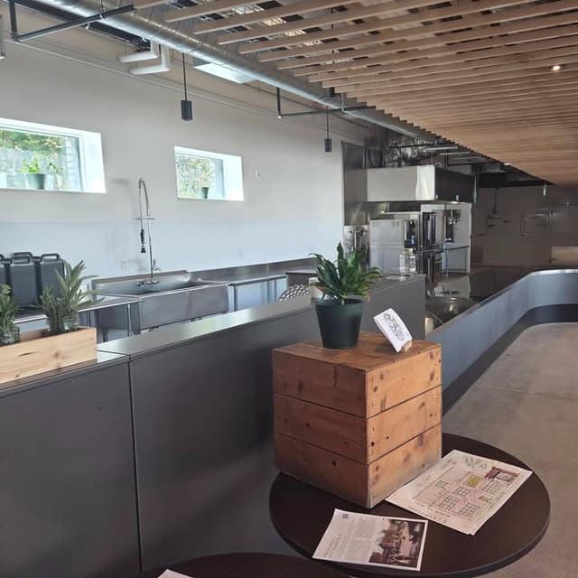 A large kitchen with stainless steel counters, a wood slat ceiling, and a small table holding a plant, a wooden crate and printed papers.