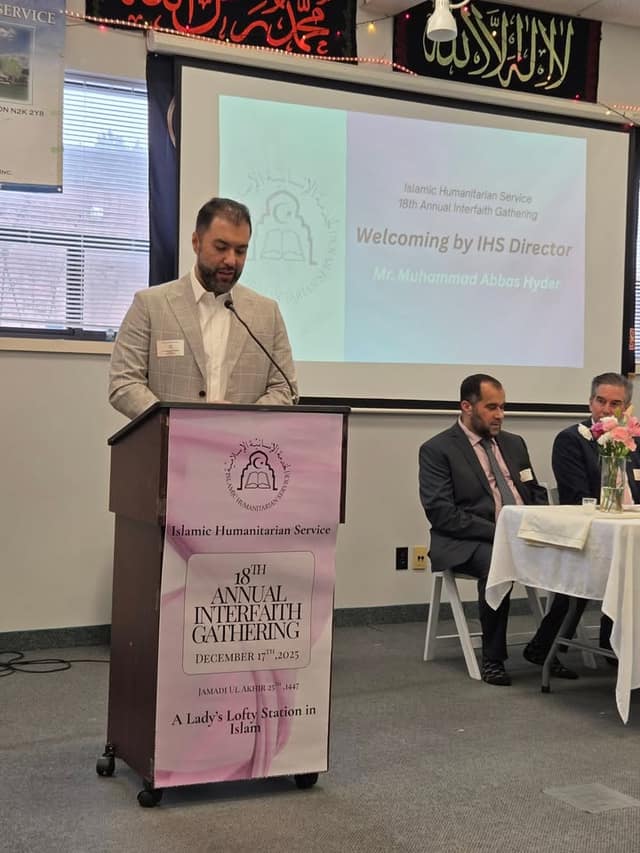 A man in a plaid blazer speaks at a podium reading "18th Annual Interfaith Gathering" as a screen shows a welcome by the IHS Director.