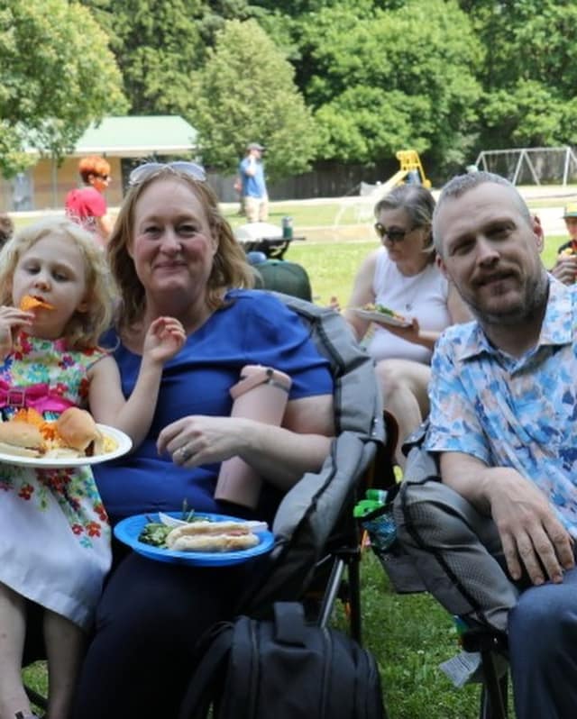 A woman in blue sits in a camp chair with plates of food beside a young girl eating chips and a smiling man in a floral shirt.