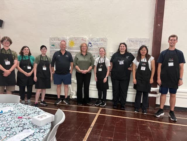Nine STEM Camp staff in black aprons and name tags stand in a row with a woman in an olive shirt and a man, in a gym with posters behind them.