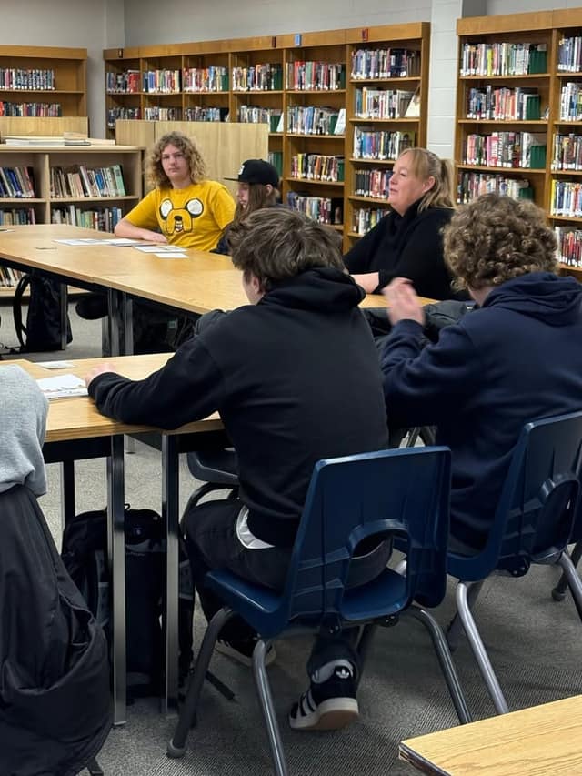 A woman in black sits at a library table across from students in hoodies, one in a yellow cartoon shirt, listening to her.