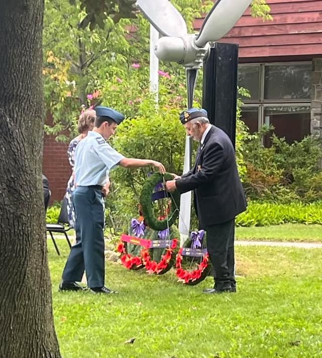 A young cadet in a light blue uniform and a veteran in a blazer and beret place poppy wreaths beside a propeller monument.