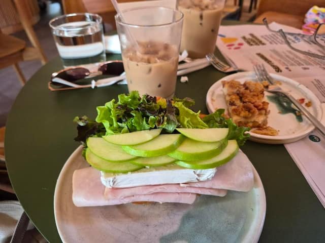 A table holds an open-faced ham and brie sandwich topped with green apple slices and lettuce, an iced latte, and a dessert.