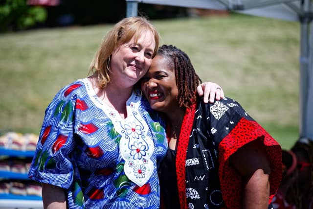 Two women embrace and smile at an outdoor event, one in a blue patterned dress, the other in a black and red robe.