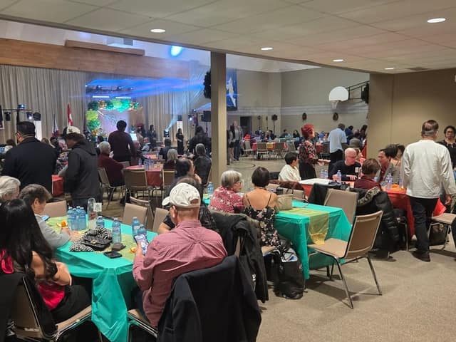 Guests sit at tables with teal and orange cloths in a banquet hall, with a decorated stage and balloon arch in the background.