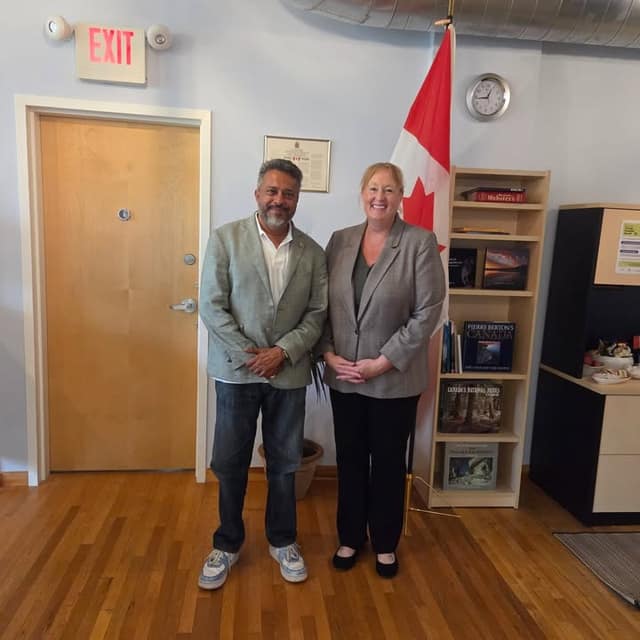 A man in a light green blazer and Kelly DeRidder stand in front of a bookshelf and Canadian flag near a wooden door.
