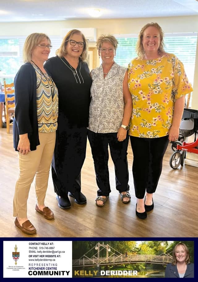 Four smiling women pose together indoors in a bright common room with wood-style flooring, with a red walker visible at right.