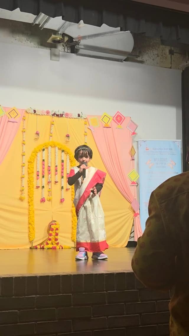 A young child in a white and red sari speaks into a microphone on stage in front of a yellow backdrop with paper kites.