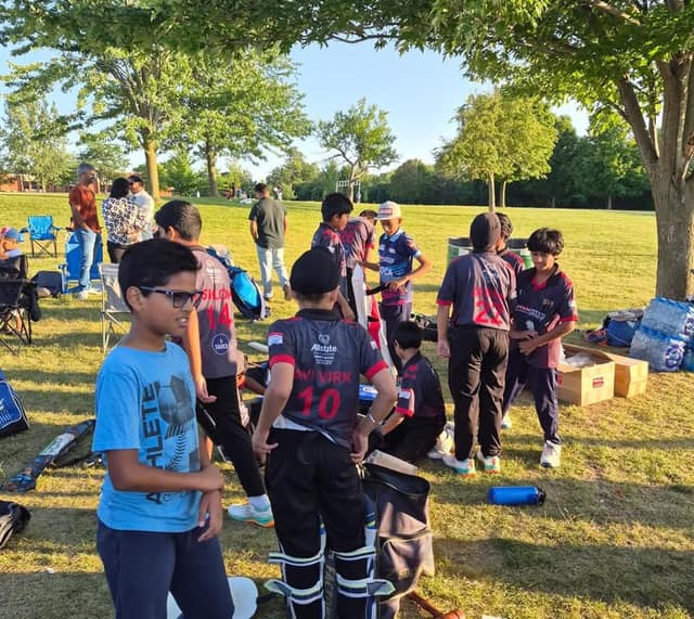 Young cricket players in jerseys gather their gear on a grassy park under trees while adults stand and talk in the background.