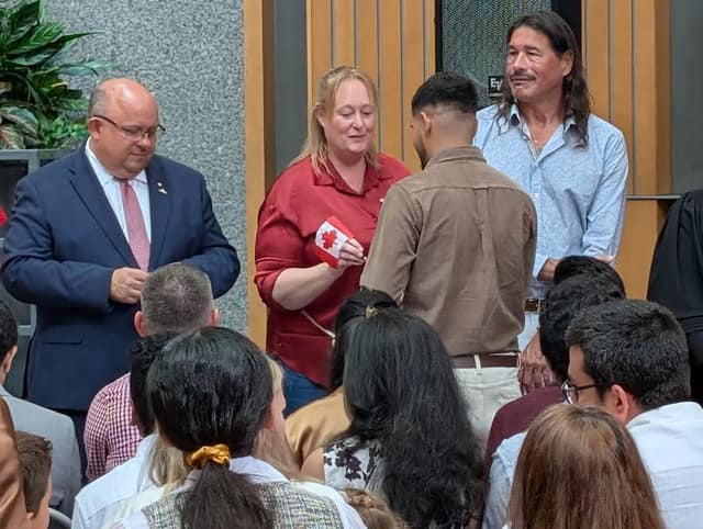 A woman in a red shirt hands a small Canadian flag to a man in a brown shirt while a man in a suit and another man look on.