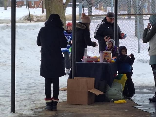 Children and adults gather around a table of supplies under a covered shelter by a fence, with snow on the ground.