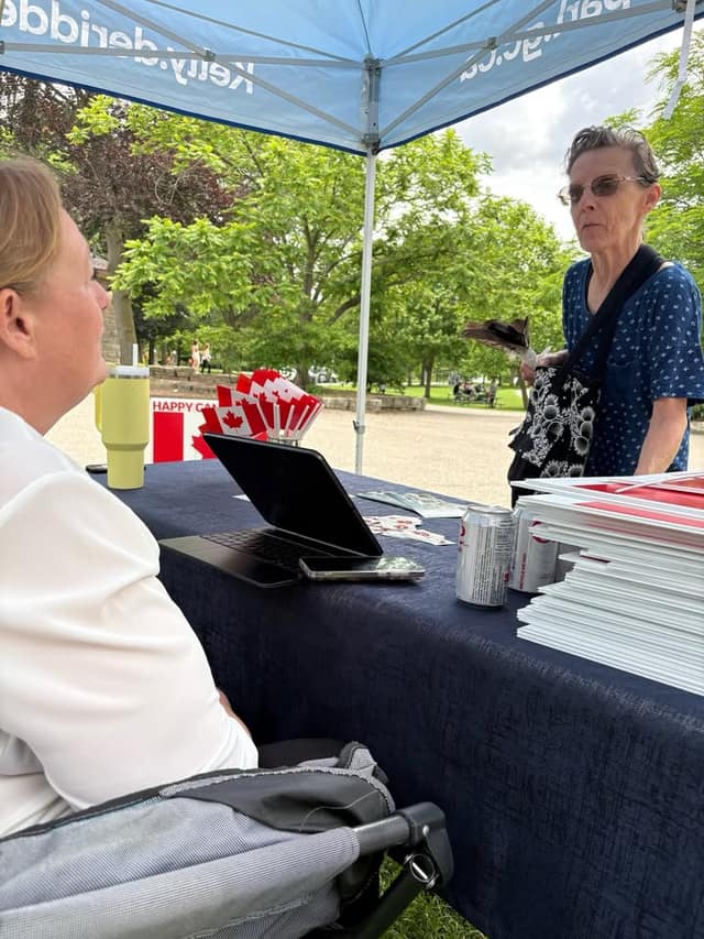 A woman in white sits at a table with a laptop, Canadian flags and lawn signs while a woman in a blue top stands nearby under the tent.