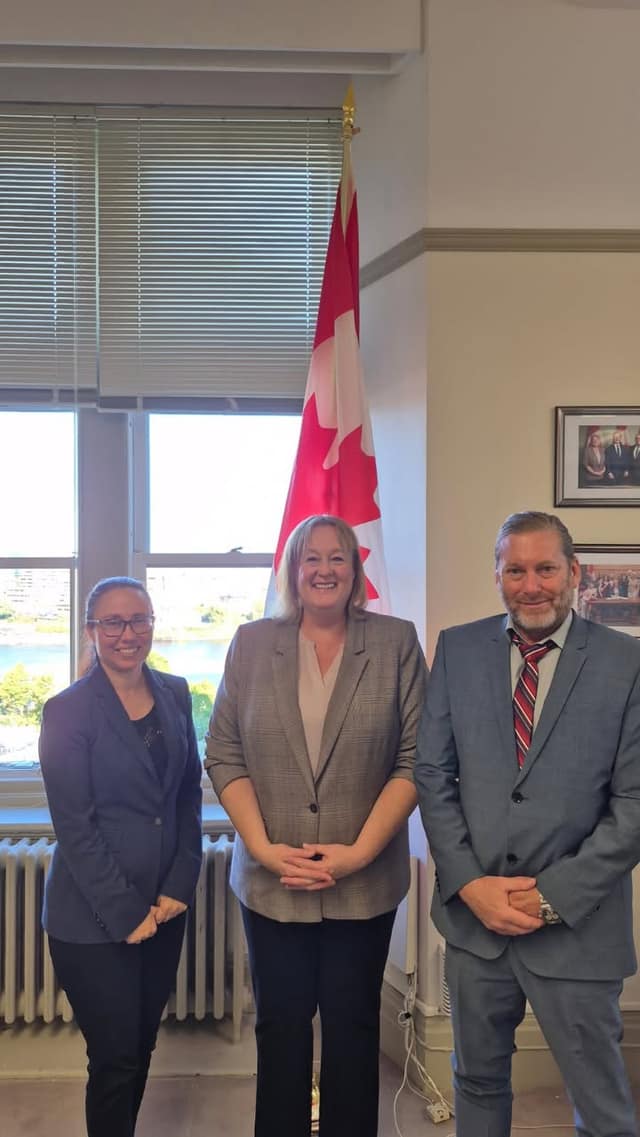 Three people in business attire stand smiling side by side in an office beside a Canadian flag, with a window and radiator behind them.
