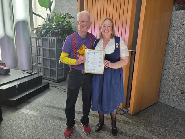 Erick Traplin, in a purple shirt and red suspenders, and a woman in a blue dirndl smile and hold a framed certificate in a lobby.