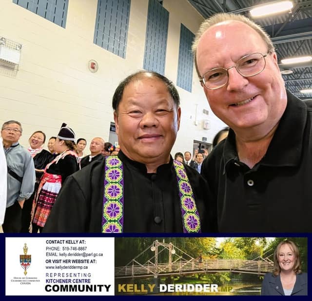 A smiling man in glasses and black polo takes a selfie with a man in a black outfit with a purple embroidered sash, in a gym with guests behind.