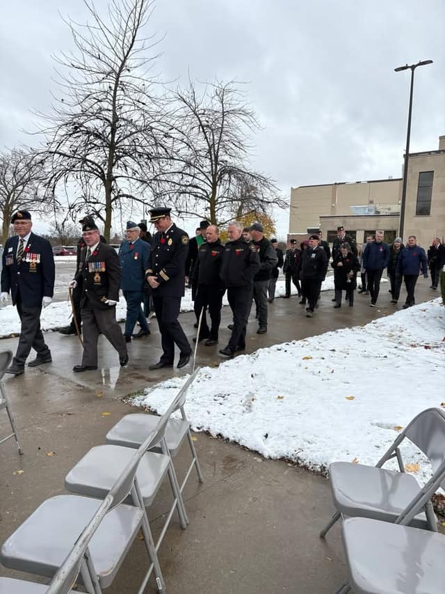 Veterans with medals, firefighters and other attendees walk past rows of empty folding chairs on a snowy, wet pavement.