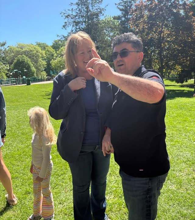A smiling woman in a blue blazer laughs beside a man in sunglasses who points upward, with a child in striped pants nearby in a park.