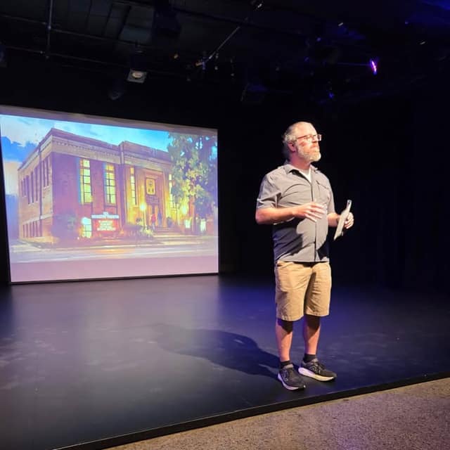 A bearded man in glasses, gray shirt and khaki shorts holds papers on a dark stage beside a projected illustration of the Registry Theatre building.