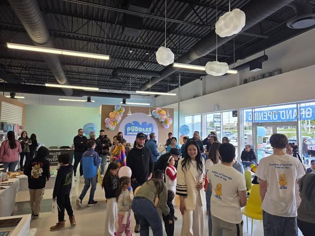 Crowd of adults and children gathers inside the bright Puddles Swim School lobby, with balloon displays and a Grand Opening sign in the window.
